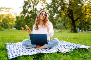 Sunny Park 'ta elinde dizüstü bilgisayarla battaniyenin üzerinde oturan serbest çalışan bir kadın. Kablosuz kulaklık takan kadın laptopta çalışıyor ve açık havada güneşli bir günün tadını çıkarıyor. Kavram, teknoloji, doğa ve rahatlama..