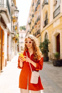 Güneşli bir caddede telefonla gezen genç bir turistin portresi. Güzel bir kadın telefona bakar, sohbet eder ve seyahat ederken güneşli bir günün tadını çıkarır. Kavramsal resim: turizm, tarz ve navigasyon.