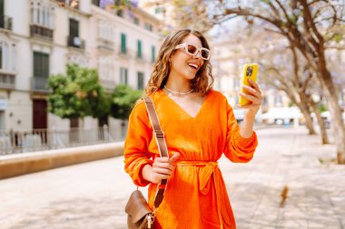 Güneşli bir günde şehir caddesinde telefonu olan turuncu elbiseli genç bir kadın. Şık bir kadın telefonuyla şehirde dolaşmaktan hoşlanır. Selfie, turizm ve moda konseptleri.