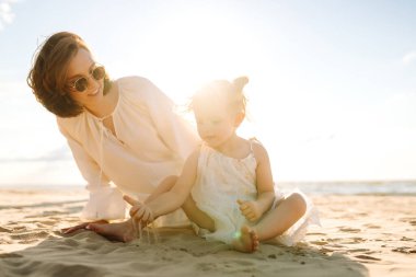 Mutlu anne ve küçük kızı sahilde birlikte eğleniyor, güneşte sarılıyorlar. Genç bir kadın ve küçük bir kız sahilde birlikte vakit geçiriyorlar. Çocukluk kavramları, annelik, rahatlama..