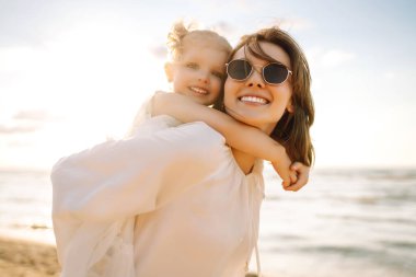 Mutlu anne ve küçük kızı sahilde birlikte eğleniyor, güneşte sarılıyorlar. Genç bir kadın ve küçük bir kız sahilde birlikte vakit geçiriyorlar. Çocukluk kavramları, annelik, rahatlama..