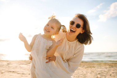 Mutlu anne ve küçük kızı sahilde birlikte eğleniyor, güneşte sarılıyorlar. Genç bir kadın ve küçük bir kız sahilde birlikte vakit geçiriyorlar. Çocukluk kavramları, annelik, rahatlama..