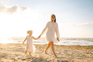 Mutlu anne ve küçük kızı sahilde birlikte eğleniyor, güneşte sarılıyorlar. Genç bir kadın ve küçük bir kız sahilde birlikte vakit geçiriyorlar. Çocukluk kavramları, annelik, rahatlama..
