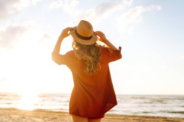 Güneşli bir günde sahilde eğlenen şapkalı genç bir kadının arka görüntüsü. Kadın kendini özgür hissediyor ve gün batımında deniz manzarasının tadını çıkarıyor. Özgürlük, rahatlama ve doğa kavramı.