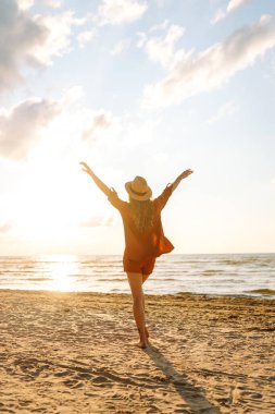 A rear view of a young woman in a hat enjoying herself on the beach on a sunny day. The woman feels free and enjoys the seascape at sunset. Concept of freedom, relaxation, and nature.