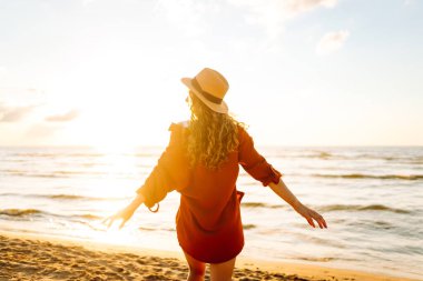 Güneşli bir günde sahilde eğlenen şapkalı genç bir kadının arka görüntüsü. Kadın kendini özgür hissediyor ve gün batımında deniz manzarasının tadını çıkarıyor. Özgürlük, rahatlama ve doğa kavramı.