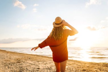 Güneşli bir günde sahilde eğlenen şapkalı genç bir kadının arka görüntüsü. Kadın kendini özgür hissediyor ve gün batımında deniz manzarasının tadını çıkarıyor. Özgürlük, rahatlama ve doğa kavramı.