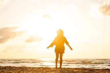 Güneşli bir günde sahilde eğlenen şapkalı genç bir kadının arka görüntüsü. Kadın kendini özgür hissediyor ve gün batımında deniz manzarasının tadını çıkarıyor. Özgürlük, rahatlama ve doğa kavramı.