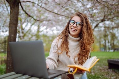 Neşeli kadın sarı defteriyle bahçedeki masada oturuyor. Genç bir kadın dizüstü bilgisayarda çalışıyor, elinde defter var, bahar günü. Serbest çalışma, rahatlama ve hafta sonları kavramı.