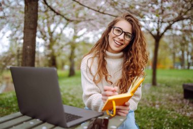 Neşeli kadın sarı defteriyle bahçedeki masada oturuyor. Genç bir kadın dizüstü bilgisayarda çalışıyor, elinde defter var, bahar günü. Serbest çalışma, rahatlama ve hafta sonları kavramı.