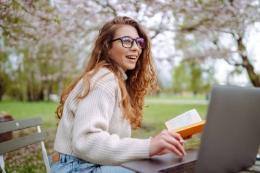 Neşeli kadın sarı defteriyle bahçedeki masada oturuyor. Genç bir kadın dizüstü bilgisayarda çalışıyor, elinde defter var, bahar günü. Serbest çalışma, rahatlama ve hafta sonları kavramı.