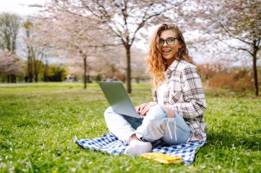 Neşeli kadın sarı defteriyle bahçedeki masada oturuyor. Genç bir kadın dizüstü bilgisayarda çalışıyor, elinde defter var, bahar günü. Serbest çalışma, rahatlama ve hafta sonları kavramı.