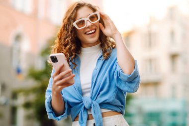 Güneşli bir caddede telefonuyla konuşan bir kadının portresi. Telefonu olan şık bir kadın dışarıda yürüyor. Blog yazma, yürüme ve turizm kavramları.