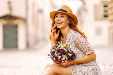 Bir buket kır çiçeğiyle mutlu kadın şehrin güneşli caddelerinde geziniyor. Şapkalı güzel bir kadın elinde bir buket çiçek tutuyor ve açık havada günbatımını seyrediyor. Kavram güzellik, eğlence ve turizmdir..