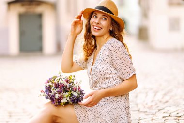 Bir buket kır çiçeğiyle mutlu kadın şehrin güneşli caddelerinde geziniyor. Şapkalı güzel bir kadın elinde bir buket çiçek tutuyor ve açık havada günbatımını seyrediyor. Kavram güzellik, eğlence ve turizmdir..