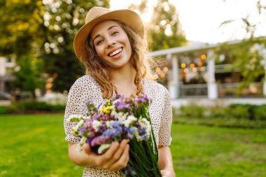 Yeşil bir çayırda bir buket kır çiçeğiyle parktaki genç bir kadının portresi. Şapkalı güzel bir kadın, elinde kır çiçekleri olan, gün batımını seyreden. Güzellik, moda ve eğlence kavramı.