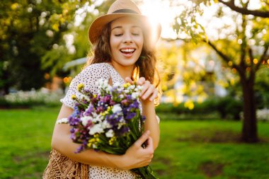 Yeşil bir çayırda bir buket kır çiçeğiyle parktaki genç bir kadının portresi. Şapkalı güzel bir kadın, elinde kır çiçekleri olan, gün batımını seyreden. Güzellik, moda ve eğlence kavramı.