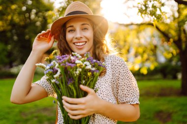 Yeşil bir çayırda bir buket kır çiçeğiyle parktaki genç bir kadının portresi. Şapkalı güzel bir kadın, elinde kır çiçekleri olan, gün batımını seyreden. Güzellik, moda ve eğlence kavramı.