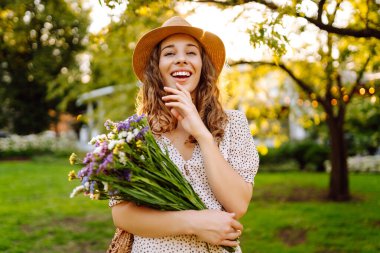Portrait of a young woman in a park with a bouquet of wildflowers in a green meadow. A beautiful woman in a hat, holding wildflowers, enjoys the sunset outdoors. Concept of beauty, fashion, and fun.