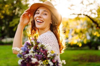 Yeşil bir çayırda bir buket kır çiçeğiyle parktaki genç bir kadının portresi. Şapkalı güzel bir kadın, elinde kır çiçekleri olan, gün batımını seyreden. Güzellik, moda ve eğlence kavramı.