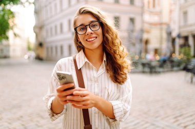 Gün batımında merkez meydanında gözlük takan ve elinde telefon tutan genç bir kadının portresi. Güneşli bir günde şık giyinmiş güzel bir kadın telefonuna mesaj atıyor. Tarz ve yürüme kavramı.