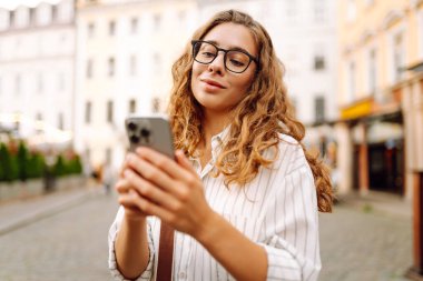 Gün batımında merkez meydanında gözlük takan ve elinde telefon tutan genç bir kadının portresi. Güneşli bir günde şık giyinmiş güzel bir kadın telefonuna mesaj atıyor. Tarz ve yürüme kavramı.