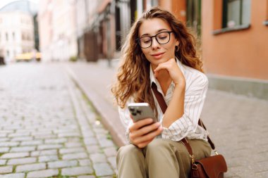 Gün batımında merkez meydanında gözlük takan ve elinde telefon tutan genç bir kadının portresi. Güneşli bir günde şık giyinmiş güzel bir kadın telefonuna mesaj atıyor. Tarz ve yürüme kavramı.