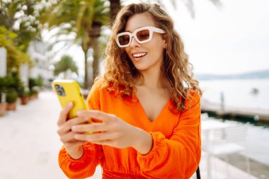 Parlak elbiseli güzel bir kadın sokakta elinde telefonla palmiye ağaçlarının yanında duruyor. Güneş gözlüklü şık bir turist güneşli toprak setinde gezintiye çıkar. Yürüme, turizm ve tarz kavramları.