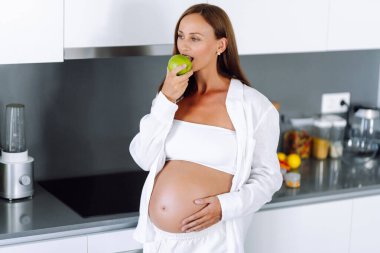 Hamile bir kadın, elinde yeşil bir elmayla modern bir mutfakta duruyor. Bebek bekleyen genç bir kadın sıcak bir odada sağlıklı bir yemek hazırlar. Beslenme, sağlık ve beklenti kavramı.