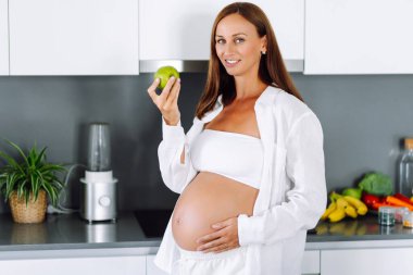 Hamile bir kadın, elinde yeşil bir elmayla modern bir mutfakta duruyor. Bebek bekleyen genç bir kadın sıcak bir odada sağlıklı bir yemek hazırlar. Beslenme, sağlık ve beklenti kavramı.