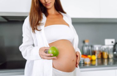 Hamile bir kadın, elinde yeşil bir elmayla modern bir mutfakta duruyor. Bebek bekleyen genç bir kadın sıcak bir odada sağlıklı bir yemek hazırlar. Beslenme, sağlık ve beklenti kavramı.
