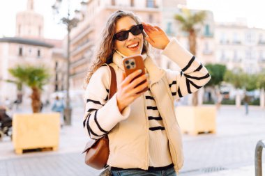 Cheerful woman wearing sunglasses and using phone strolls along sunny streets of city. Tourist admires  city's architecture in rays of the setting sun. Concepts: tourism, technology, weekends.