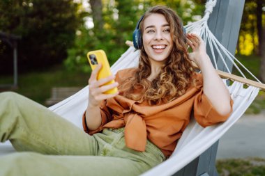 Kablosuz kulaklıkları ve telefonu olan neşeli bir kadın güneşli bir parkta hamakta dinleniyor. Genç kadın müzik dinliyor, elinde parlak bir çantayla telefon tutuyor. Rahatlama ve hafta sonu kavramı.