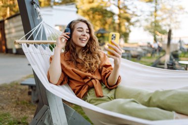 Kablosuz kulaklıkları ve telefonu olan neşeli bir kadın güneşli bir parkta hamakta dinleniyor. Genç kadın müzik dinliyor, elinde parlak bir çantayla telefon tutuyor. Rahatlama ve hafta sonu kavramı.