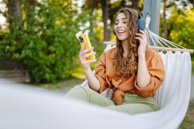 Kablosuz kulaklıkları ve telefonu olan neşeli bir kadın güneşli bir parkta hamakta dinleniyor. Genç kadın müzik dinliyor, elinde parlak bir çantayla telefon tutuyor. Rahatlama ve hafta sonu kavramı.