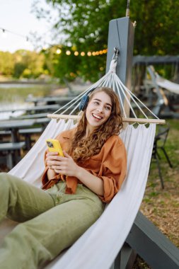 Kablosuz kulaklıkları ve telefonu olan neşeli bir kadın güneşli bir parkta hamakta dinleniyor. Genç kadın müzik dinliyor, elinde parlak bir çantayla telefon tutuyor. Rahatlama ve hafta sonu kavramı.