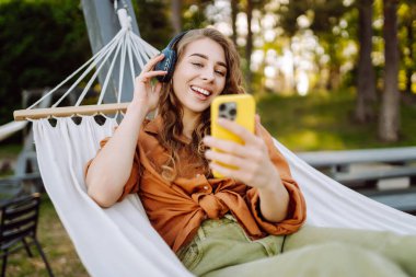 Kablosuz kulaklıkları ve telefonu olan neşeli bir kadın güneşli bir parkta hamakta dinleniyor. Genç kadın müzik dinliyor, elinde parlak bir çantayla telefon tutuyor. Rahatlama ve hafta sonu kavramı.