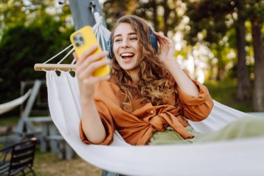 Kablosuz kulaklıkları ve telefonu olan neşeli bir kadın güneşli bir parkta hamakta dinleniyor. Genç kadın müzik dinliyor, elinde parlak bir çantayla telefon tutuyor. Rahatlama ve hafta sonu kavramı.