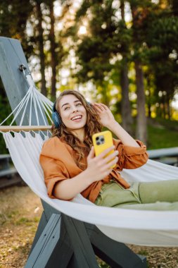 Kablosuz kulaklıkları ve telefonu olan neşeli bir kadın güneşli bir parkta hamakta dinleniyor. Genç kadın müzik dinliyor, elinde parlak bir çantayla telefon tutuyor. Rahatlama ve hafta sonu kavramı.