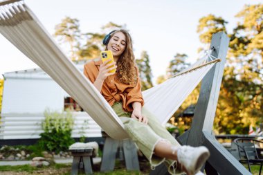 Kablosuz kulaklıkları ve telefonu olan neşeli bir kadın güneşli bir parkta hamakta dinleniyor. Genç kadın müzik dinliyor, elinde parlak bir çantayla telefon tutuyor. Rahatlama ve hafta sonu kavramı.