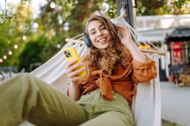 Kablosuz kulaklıkları ve telefonu olan neşeli bir kadın güneşli bir parkta hamakta dinleniyor. Genç kadın müzik dinliyor, elinde parlak bir çantayla telefon tutuyor. Rahatlama ve hafta sonu kavramı.