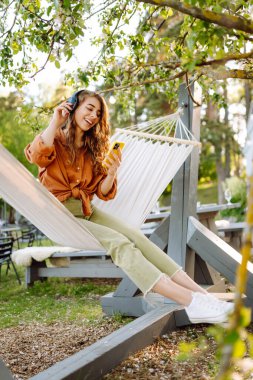Kablosuz kulaklıkları ve telefonu olan neşeli bir kadın güneşli bir parkta hamakta dinleniyor. Genç kadın müzik dinliyor, elinde parlak bir çantayla telefon tutuyor. Rahatlama ve hafta sonu kavramı.