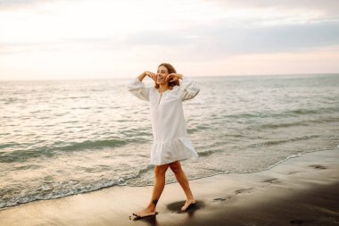 Beyaz elbiseli güzel bir kadın gün batımında deniz kıyısında geziniyor. Genç kadın deniz manzarasının tadını çıkarıyor ve kendini özgür hissediyor. Rahatlama, hafta sonları ve keyif kavramları.