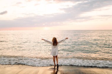 Beyaz elbiseli güzel bir kadın gün batımında deniz kıyısında geziniyor. Genç kadın deniz manzarasının tadını çıkarıyor ve kendini özgür hissediyor. Rahatlama, hafta sonları ve keyif kavramları.