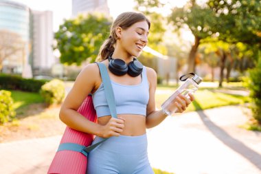 Kablosuz kulaklıkları ve yoga minderiyle atletik kadın güneşli parkta su içiyor. Genç bir kadın idmandan sonra susuzluğunu giderir ve gün batımında müzik dinlemekten hoşlanır. Spor ve rahatlama kavramı.