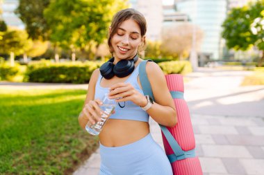 Kablosuz kulaklıkları ve yoga minderiyle atletik kadın güneşli parkta su içiyor. Genç bir kadın idmandan sonra susuzluğunu giderir ve gün batımında müzik dinlemekten hoşlanır. Spor ve rahatlama kavramı.