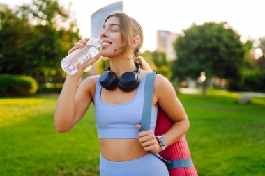 Kablosuz kulaklıkları ve yoga minderiyle atletik kadın güneşli parkta su içiyor. Genç bir kadın idmandan sonra susuzluğunu giderir ve gün batımında müzik dinlemekten hoşlanır. Spor ve rahatlama kavramı.