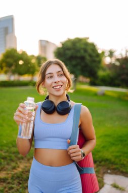 Kablosuz kulaklıkları ve yoga minderiyle atletik kadın güneşli parkta su içiyor. Genç bir kadın idmandan sonra susuzluğunu giderir ve gün batımında müzik dinlemekten hoşlanır. Spor ve rahatlama kavramı.
