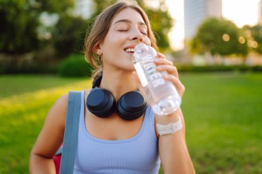 Kablosuz kulaklıkları ve yoga minderiyle atletik kadın güneşli parkta su içiyor. Genç bir kadın idmandan sonra susuzluğunu giderir ve gün batımında müzik dinlemekten hoşlanır. Spor ve rahatlama kavramı.