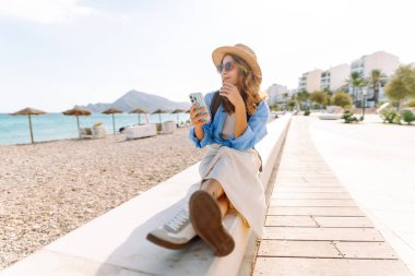 Şapka takan ve sırt çantası taşıyan bir gezgin kumsalın yanındaki bankta oturmuş, elinde telefon tutuyordu. Telefonu olan genç bir kadın gün batımında deniz manzarasının tadını çıkarıyor. Turizm, eğlence ve doğa için kavramsal fikirler.